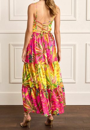 Nainen, joka pukeutunut värikkääseen, kerrokselliseen maksi-mekkoon, jossa on trooppinen kuvio, ristikkäiset avoimet selkäosat ja ohuet olkaimet, seisoo puulattialla.