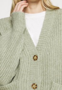 Světlě zelený houpavý pletený cardigan s knoflíky ve tvaru želvoviny, nošený přes bílý tričko, ruka se dotýká obličeje poblíž brady.