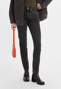 Persona vistiendo jeans ajustados negros, una camisa a rayas y una chaqueta de mezclilla negra, sosteniendo un pequeño bolso rojo, de pie contra un fondo blanco.