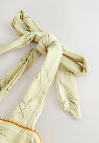 Halvány sárga sál díszes dőlt betűs írással, lazán megkötve masnira, egyik szélén arany gyöngyökkel díszítve.