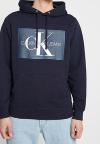 Tmavě modrá mikina s kapucí a přední kapsou na šířku, která nese velké bílé logo „CK“ a nápis „CALVIN KLEIN JEANS“ na pozadí připomínajícím džínovinu.