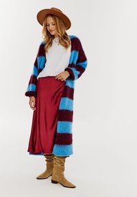 Pasiasty kardigan w niebiesko-burgundowym kolorze, dzianinowa tekstura, zestawiony z białą bluzką i czerwoną satynową spódnicą. Brązowy kapelusz z szerokim rondem i beżowe botki.