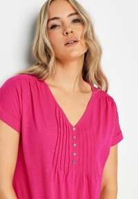 Top fucsia con scollo a V, maniche corte, pieghe verticali e una patta con sette bottoni decorativi. Texture morbida, design casual.