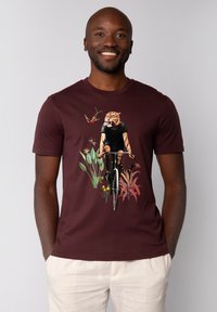 Bordeaux kleurig t-shirt met korte mouwen, versierd met een afbeelding van een tijger op een fiets, omringd door planten en vogels. Gladde katoenen stof, casual pasvorm.