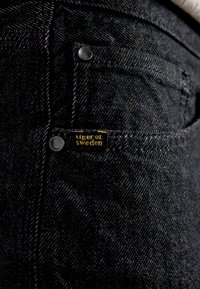 Černé džíny ze špinavé denim tkaniny s texturovaným povrchem, na kapse je žlutý štítek značky "Tiger of Sweden" a kovové nýty.