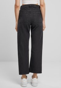 Jeans negros de tiro alto y pernera ancha con un ligero brillo, que cuentan con dos bolsillos traseros y se combinan con zapatillas blancas. Fondo liso.