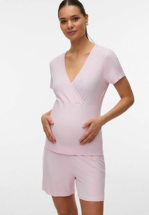 Zwangere vrouw draagt lichtroze geribd T-shirt met korte mouwen en bijpassende korte broek, houdt zachtjes haar buik vast, staand tegen een witte achtergrond.