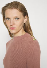 Woman with light brown hair and green eyes wearing a mauve ribbed sweater with two gold buttons on the shoulder and small hoop earrings.