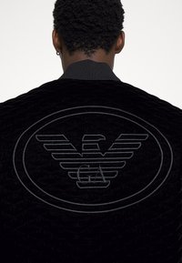 Svart quiltad jacka med en upphöjd, präglad logotyp av vingar och "GA" på baksidan, med ribbad krage och texturerad tyg.