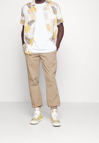 Kortärmad skjorta med palmiga mönster i beige och gult över en vit t-shirt, ihop med beiga byxor och högskor med gula accenter.