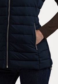 Mornarska pufasta prsluk s horizontalnim šavovima, džepovima na zip, i srebrnim detaljima. Uparen s smešnim dugim rukavima ispod.
