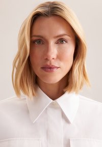 Camicia bianca con bottoni, caratterizzata da un colletto classico, due tasche frontali e una texture di tessuto liscia. Il modello ha i capelli biondi fino alle spalle.