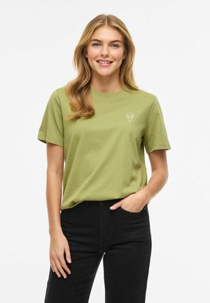 Femeie zâmbitoare cu păr lung și blond, purtând un tricou verde deschis cu un mic logo alb în formă de cornet de înghețată și pantaloni negri.