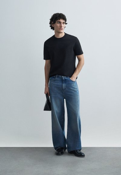 Μαύρο κοντομάνικο μπλουζάκι συνδυασμένο με μπλε τζιν παντελόνι φαρδιάς γραμμής. Το μοντέλο κρατάει μια μαύρη τσάντα και φορά μαύρα παπούτσια, στέκεται μπροστά από φωτεινό φόντο.