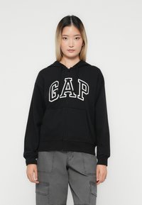 Kvinne iført svart hettejakke med "GAP" logo og grå cargo-bukser, står foran en helt hvit bakgrunn.