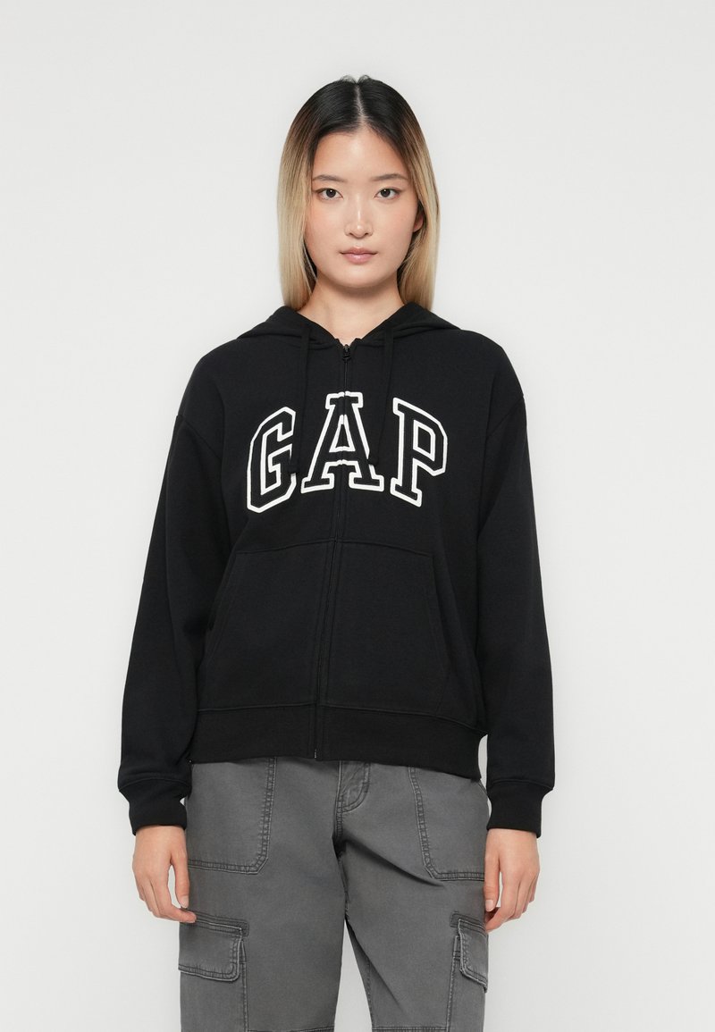 Kvinne iført svart hettejakke med "GAP" logo og grå cargo-bukser, står foran en helt hvit bakgrunn.