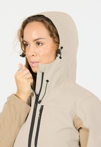 Vrouw in een beige jas met capuchon, die de ritssluiting bij haar kin aanpast, met een neutrale achtergrond.