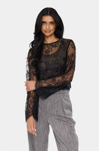 Fekete csipkeblúz hosszú ujjakkal, virágmintával, áttetsző textúrával és kerek nyakkivágással. Szürke csíkos nadrággal párosítva.