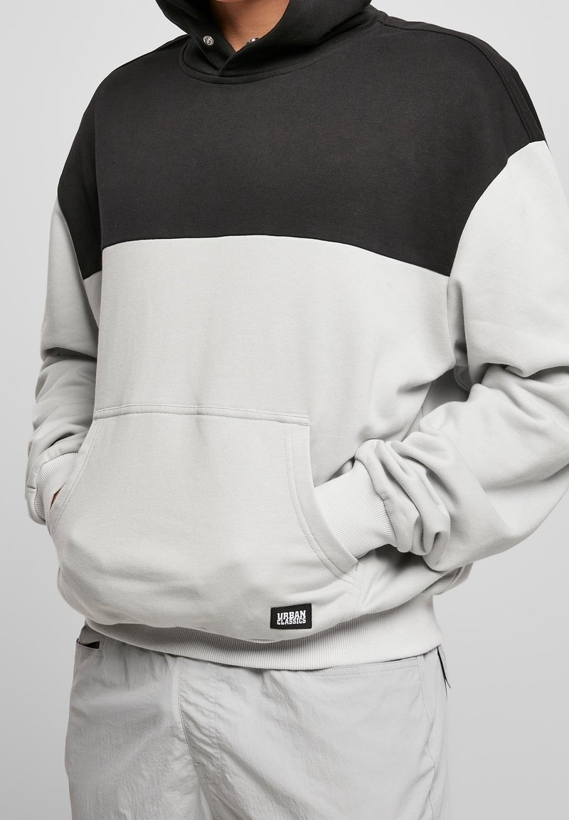 Sweat à capuche gris clair et noir avec une poche kangourou à l'avant, des poignets côtelés et un écusson logo à l'ourlet. Tissu doux et coupe décontractée.