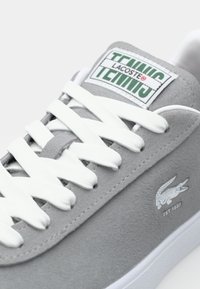 Grijze suède sneaker met witte veters, een groen en witte 'Tennis' label, en een krokodillogo. Zachte textuur met een minimalistisch ontwerp.