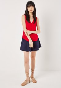 Red sleeveless top with button-down front and V-neck, paired with a navy skirt. Tan strappy heels complete the outfit. Smooth fabric texture.