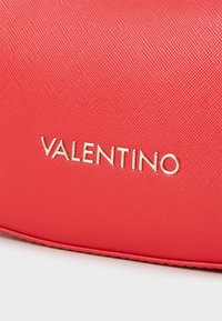 Sarkans teksturēts auduma maisiņš ar metāliski zelta "VALENTINO" logo priekšpusē. Gluda apdare ar smalkiem rakstiem un šuvju detaļām.