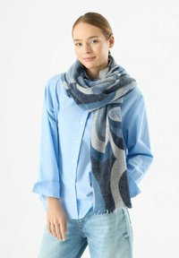 Sciarpa a fantasia blu e grigia drappeggiata attorno al collo, realizzata in materiale morbido con bordi frangiati, abbinata a una camicia blu chiaro con bottoni.