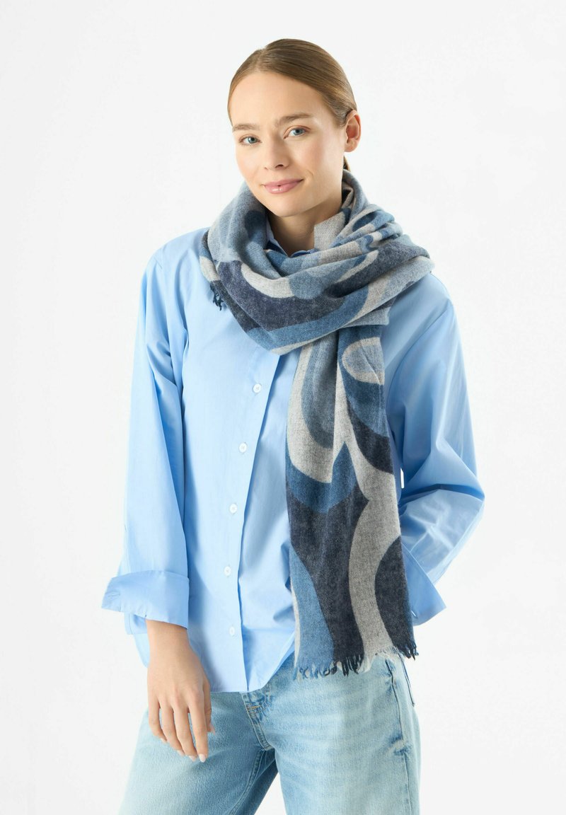 Sciarpa a fantasia blu e grigia drappeggiata attorno al collo, realizzata in materiale morbido con bordi frangiati, abbinata a una camicia blu chiaro con bottoni.
