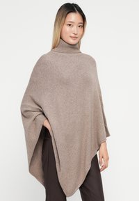 Poncho castanho claro com gola alta, feito de um tecido de malha suave com uma textura lisa e um formato fluido, que se drapeia sobre o corpo.