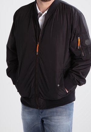 Homme portant une veste bomber noire avec des tirettes de fermeture éclair orange, une chemise blanche en dessous, et un jean bleu, les mains dans les poches de la veste.