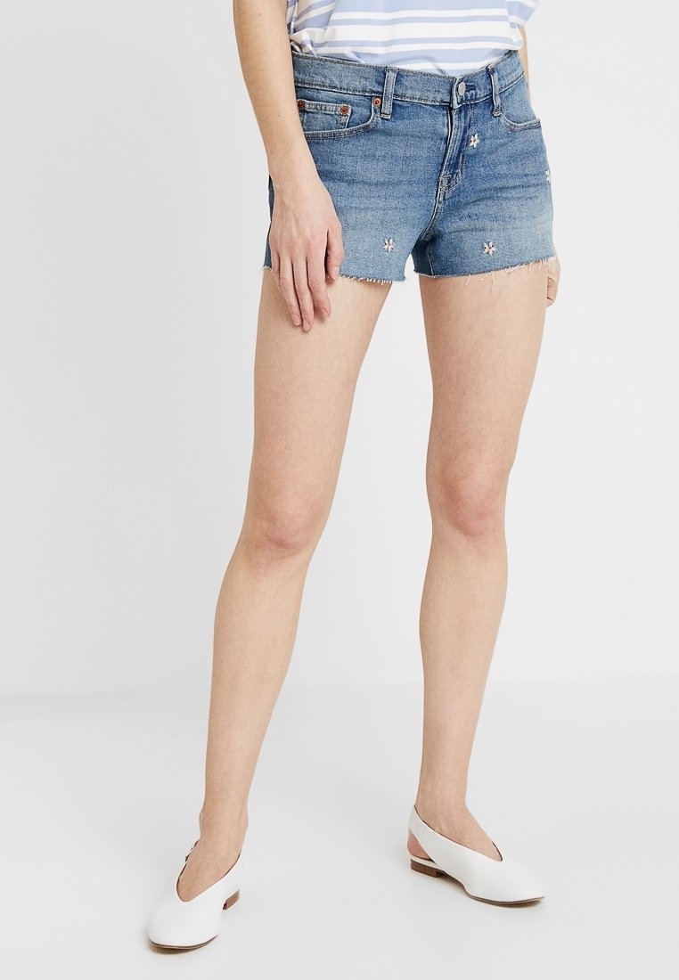 Shorts de mezclilla en azul pastel con un dobladillo deshilachado, que presentan detalles florales bordados. Diseño clásico de cinco bolsillos, ajuste a mitad de cintura.