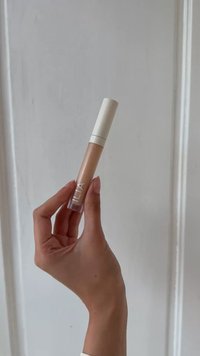 Lipgloss in een doorzichtige tube met een witte dop, met een gladde, lichtbeige formule. Etiket zegt "ILIA." Hand die het product vasthoudt.