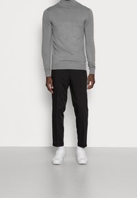Homme portant un pull-over en col roulé gris, un pantalon noir sur mesure, des chaussettes blanches et des baskets blanches, se tenant contre un mur blanc uni.