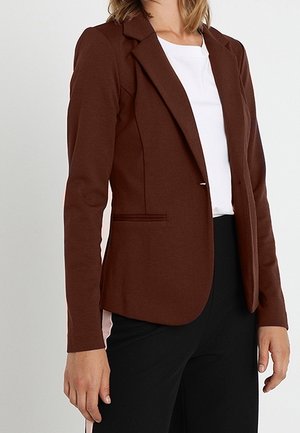 Femme portant un blazer marron cintré sur une chemise blanche et un pantalon noir avec une bande beige sur le côté, debout les bras détendus.