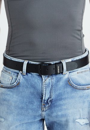 Svart läderbälte med en rektangulär spänne, bärs över urtvättade blå jeans. Jeansen har en sliten textur och synliga sömmar.