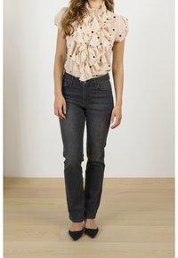 Beige blouse met ruffles en zwarte stippen, gecombineerd met donker denim jeans en zwarte puntige hakken, tegen een lichte achtergrond.
