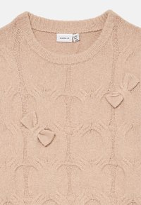 Maglione lavorato a maglia beige chiaro con un motivo circolare, caratterizzato da tre fiocchi decorativi. Il collo rotondo a costine aggiunge texture. Materiale morbido.