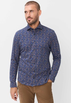 Mand med kort hår og skæg iført en blå blomsterprint langærmet skjorte og brune bukser, stående med den ene hånd i lommen.