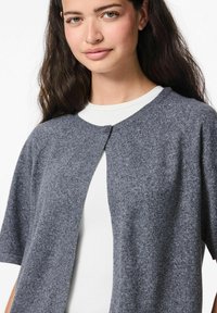 Tumši pelēks īsām piedurknēm, atvērtas priekšpuses kardigāns, izgatavots no mīksta teksturēta materiāla. Iekļauj vienu pogu un apaļu kakla izgriezumu.