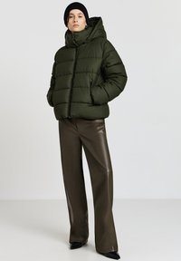 Giacca imbottita colore verde oliva con collo alto e cappuccio, chiusura con zip, dotata di tasche laterali. Indossata con pantaloni marroni lucidi a vita alta.