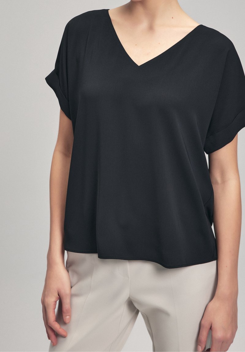 Blouse noire à col en V avec manches courtes retroussées, en tissu lisse, présentant une coupe ample qui tombe doucement sur le corps.