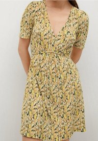 Vestido floral con escote en V, mangas cortas abullonadas y cintura ajustada. Presenta un patrón floral en amarillo, blanco y verde sobre un fondo claro.