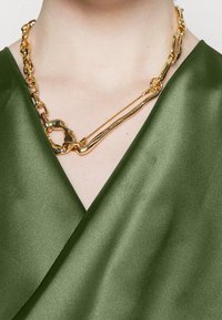 Collier en chaîne dorée avec des maillons entrelacés ; porté sur un haut en satin vert foncé avec un design en V et une texture lisse.