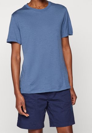 Person wearing a plain short-sleeve blue t-shirt and dark blue knee-length shorts against a plain light background.