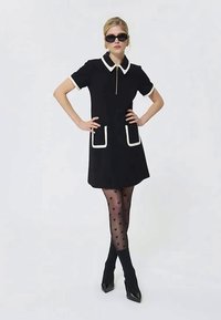 Rochie scurtă neagră cu guler alb și accente de buzunare; modelul poartă ciorapi modelați cu inimioare și pantofi cu toc. Ochelari de soare incluși.