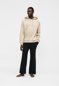 Homme portant un sweat à capuche beige avec texte et un pantalon large noir, debout pieds nus devant un fond clair uni.