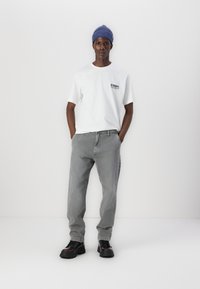 Λευκή κοντομάνικη μπλούζα με μαύρο λογότυπο, γκρι φαρδιά τζιν παντελόνια και μαύρα ογκώδη παπούτσια. Μπλε ριμπ σκουφάκι στο κεφάλι.