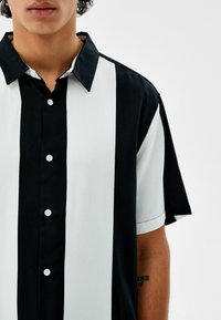 Musta-valkoraidallinen, lyhythihainen paita, jossa on napillinen kiinnitys, perinteinen kaulus ja sileä, kudottu pinta.