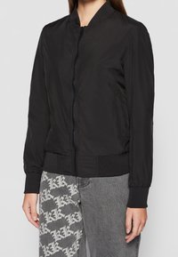 Veste bomber noire avec une finition lisse et mate. Elle est dotée d'une fermeture éclair à l'avant, de poignets et d'une ceinture côtelés, avec une silhouette ajustée.