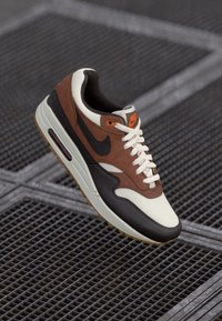 Sapatilha Nike castanha, preta e branca com atacadores brancos suspensa sobre um piso em grelha de metal.